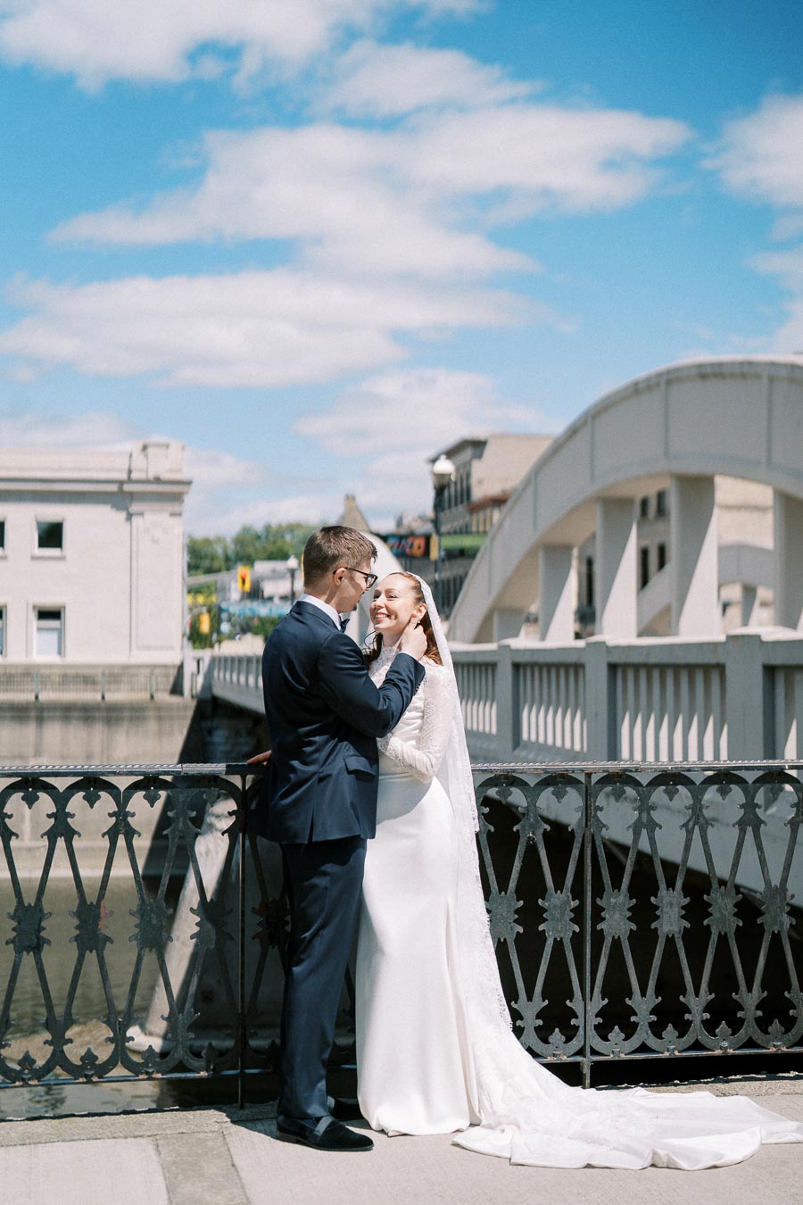 Wedding couple embracing on a bridge under a bright blue sky, wearing elegant attire, with an ornate railing and white building in the background.