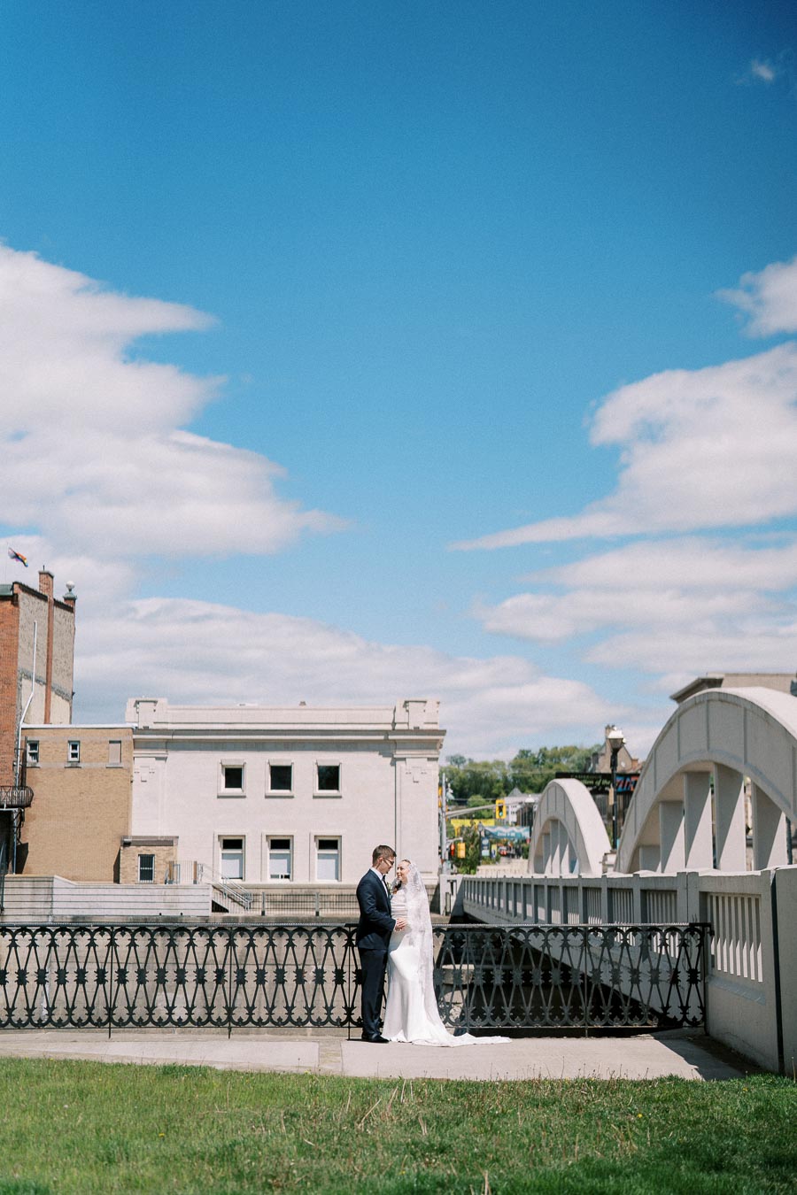 A couple in wedding attire stands on an outdoor bridge under a clear blue sky, surrounded by urban architecture.