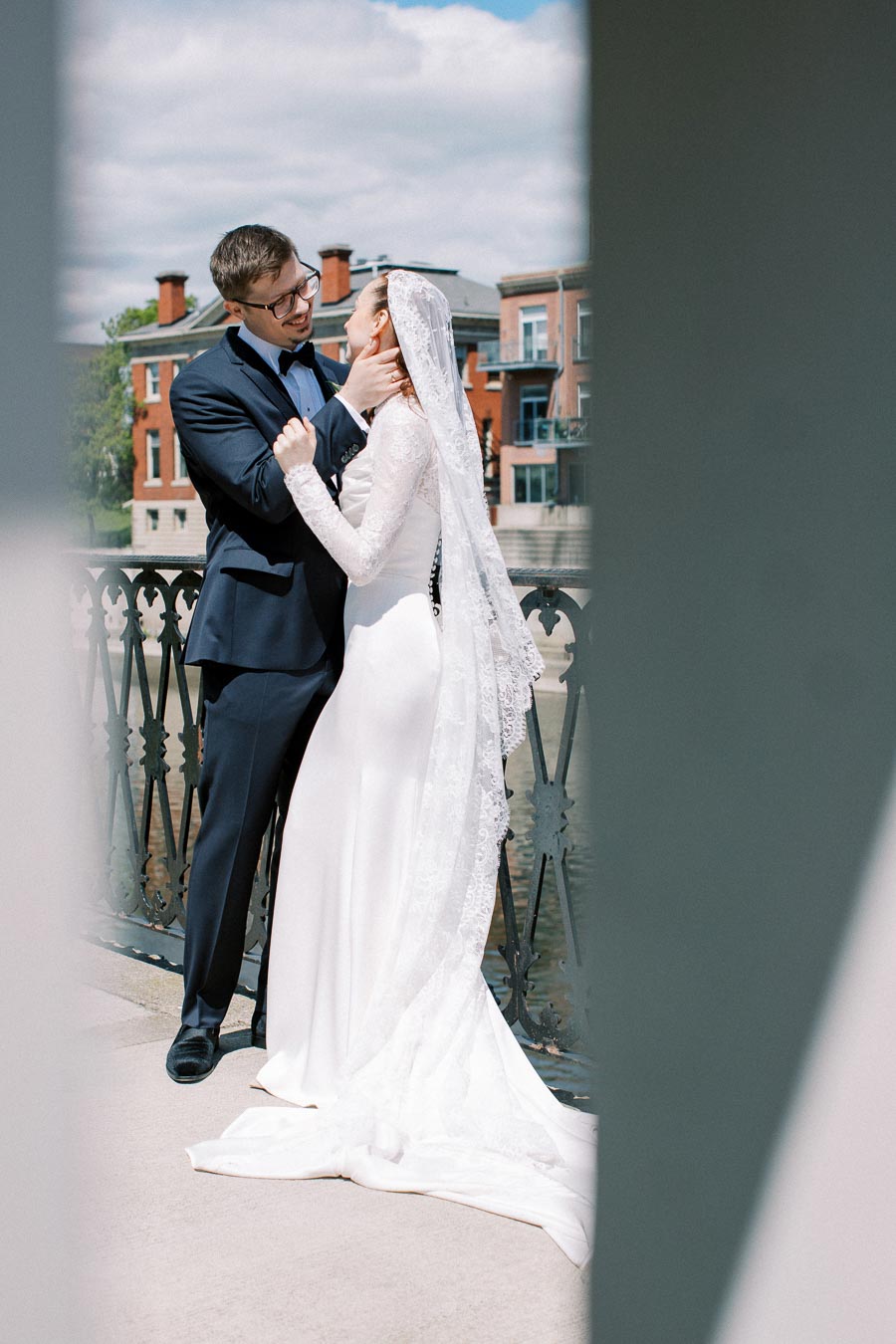 A bride in an elegant white dress and veil embraces a groom in a navy suit on a bridge, with a backdrop of charming brick buildings and a cloudy sky.