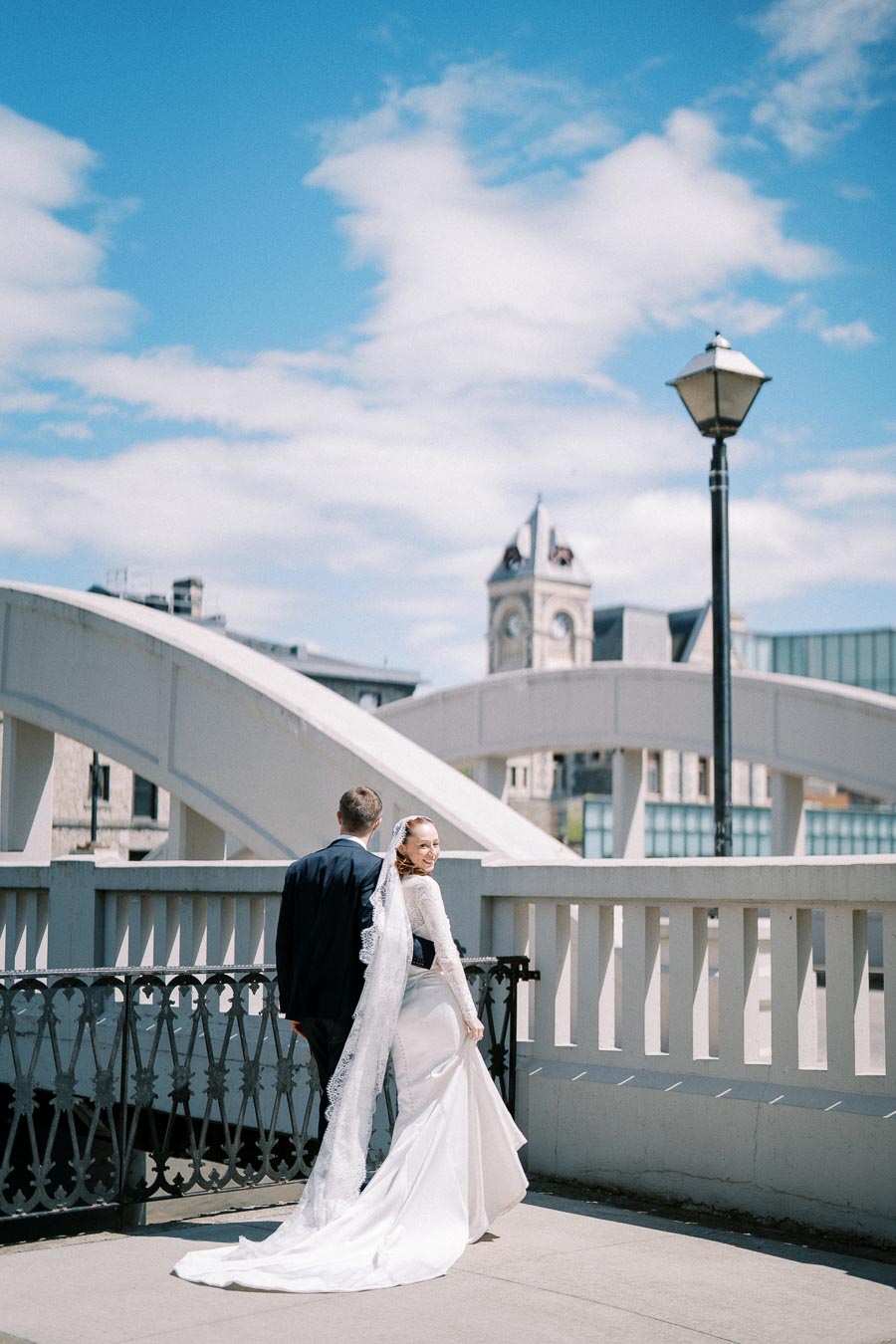 Wedding couple on a bridge with stunning architecture and blue sky backdrop, bride in white gown and groom in black suit, romantic outdoor setting.