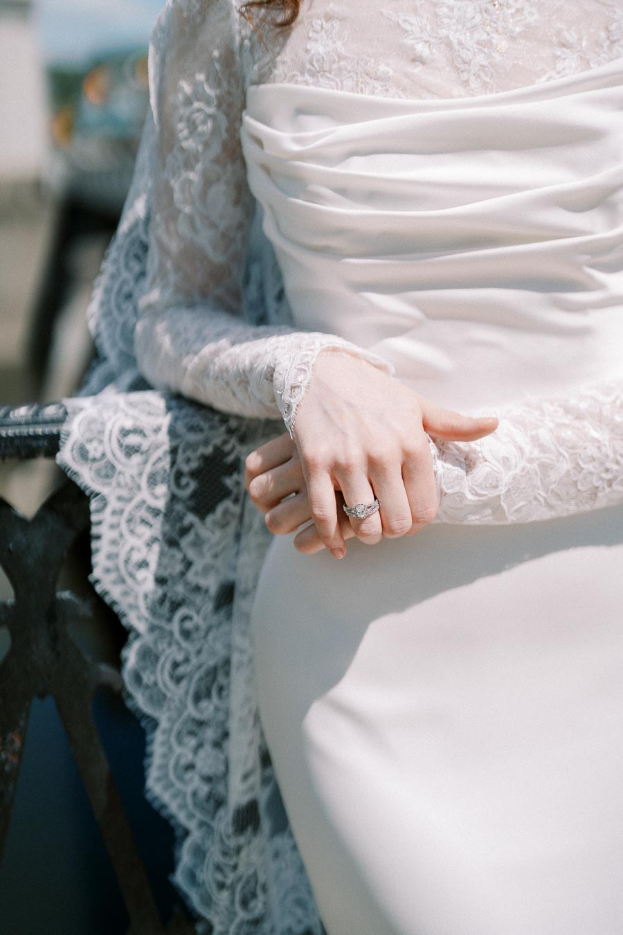 A close-up of a bride's hands resting on a decorative railing, showcasing a detailed lace wedding dress and a sparkling engagement ring.