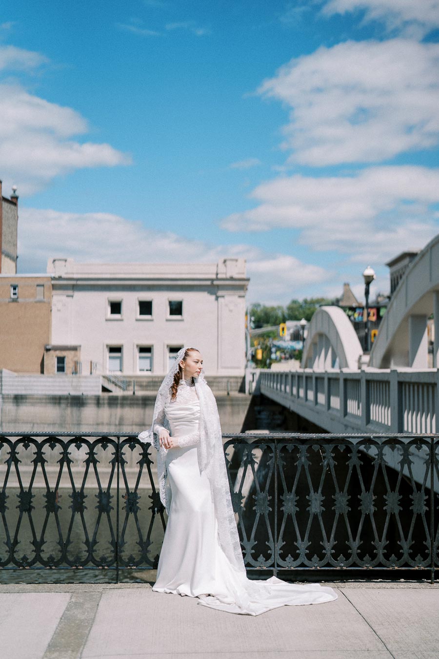 Bride in elegant white gown standing on a bridge with ornate railing, set against a backdrop of historic architecture and a bright blue sky with clouds.