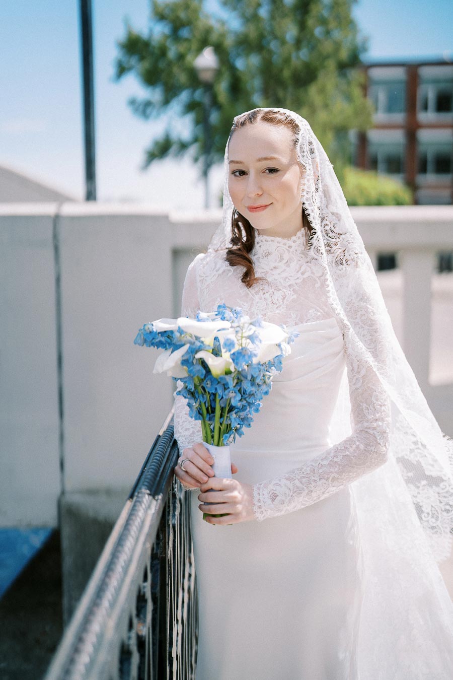A bride in a long-sleeved, lace wedding dress holding a bouquet of blue and white flowers, standing outdoors beside a railing on a sunny day.