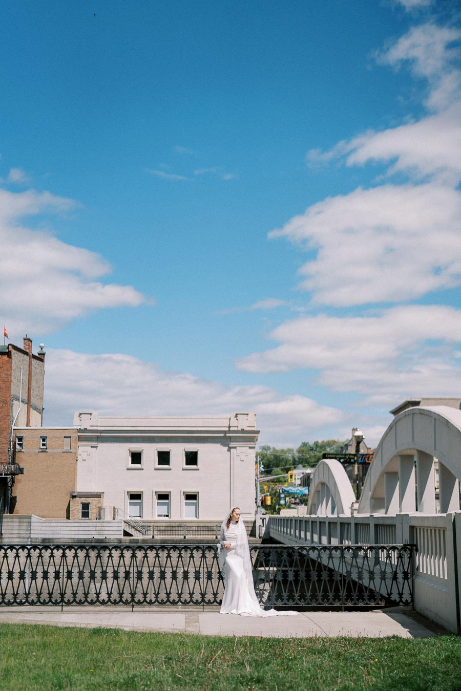 A bride in a long white dress and veil stands on a bridge, with historic buildings and a bright blue sky in the background.