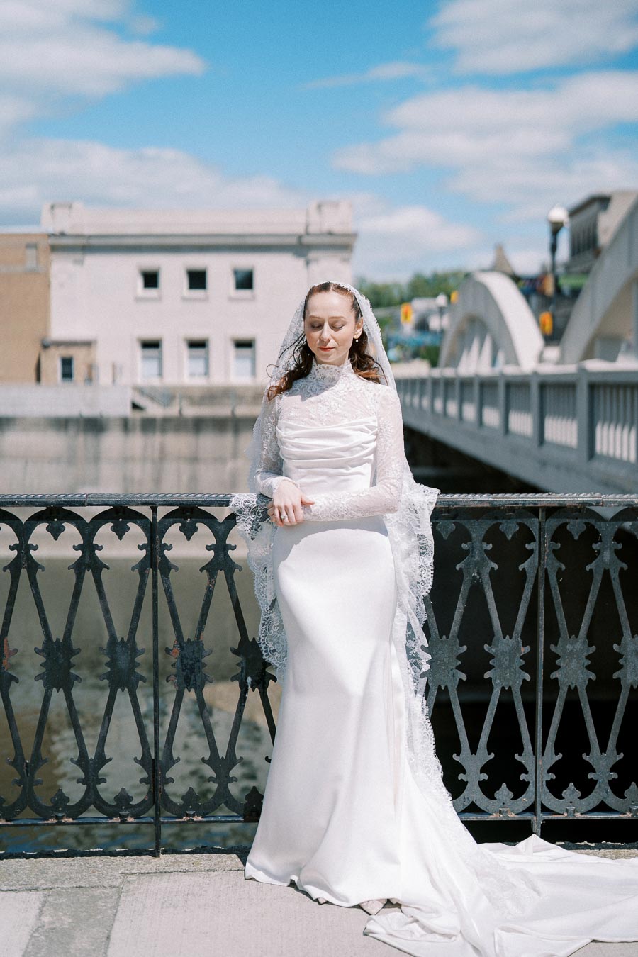 Bride in elegant white lace wedding gown posing on a bridge with ornate railing, under a bright blue sky.