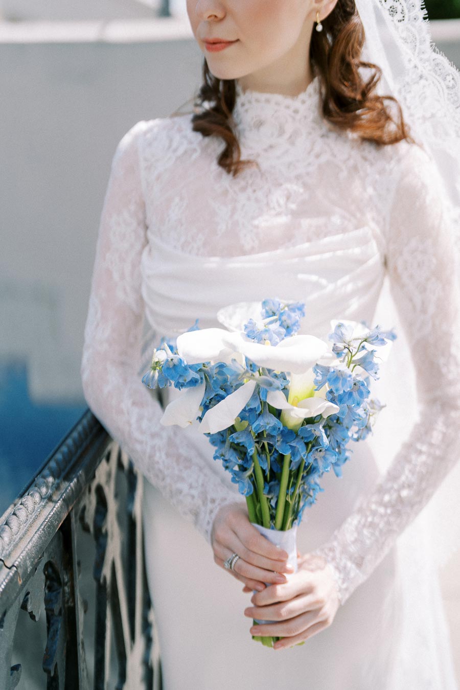 Bride in a white lace wedding dress holding a bouquet of blue and white flowers, standing by a railing.
