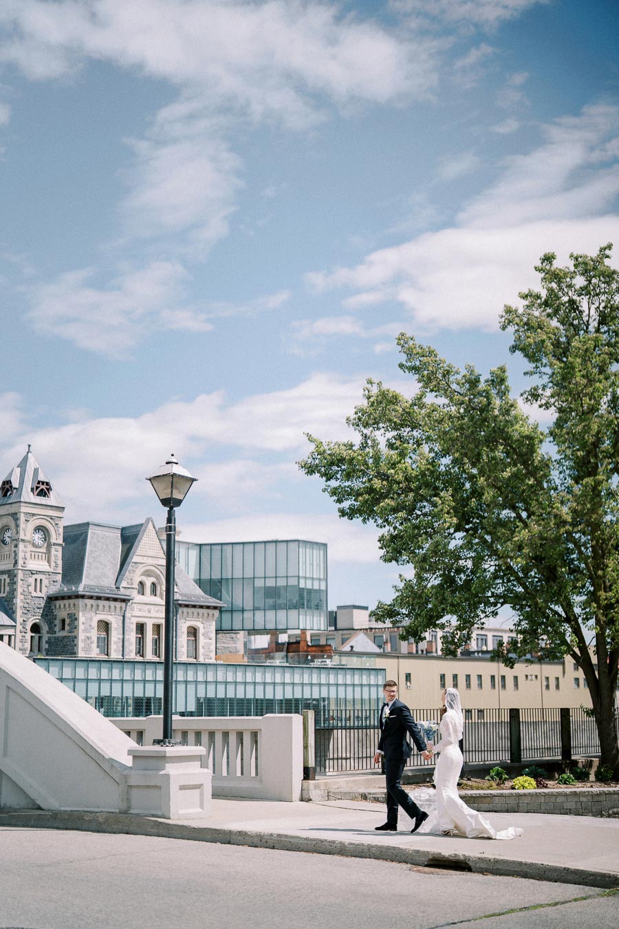 Bride and groom walking hand in hand on a sunny day, set against a backdrop of historic buildings and modern architecture, with a clear blue sky above.