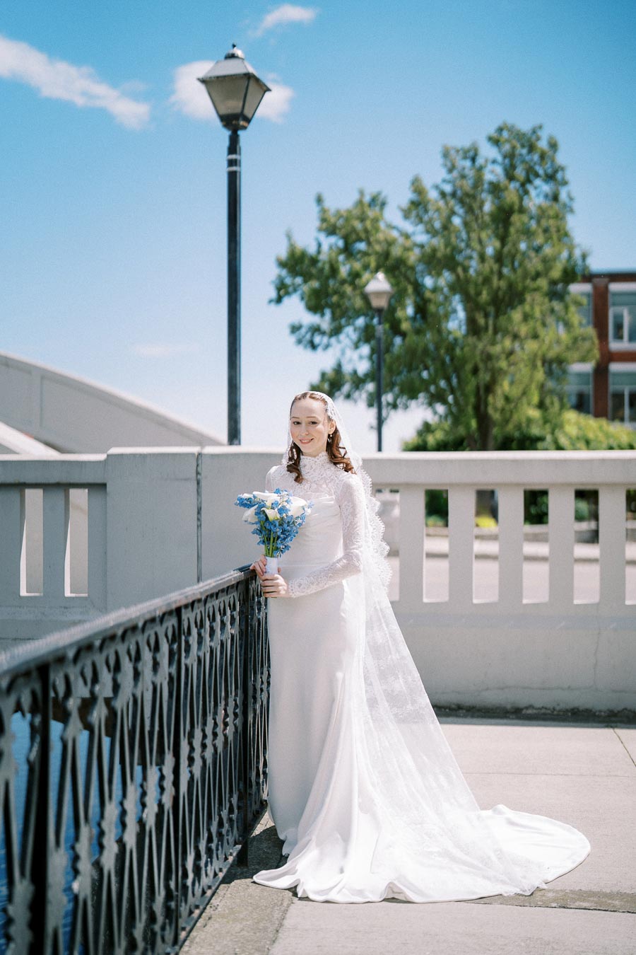 Bride in lace wedding gown holding blue bouquet stands by a railing on a sunny day, with a lamppost and trees in the background.