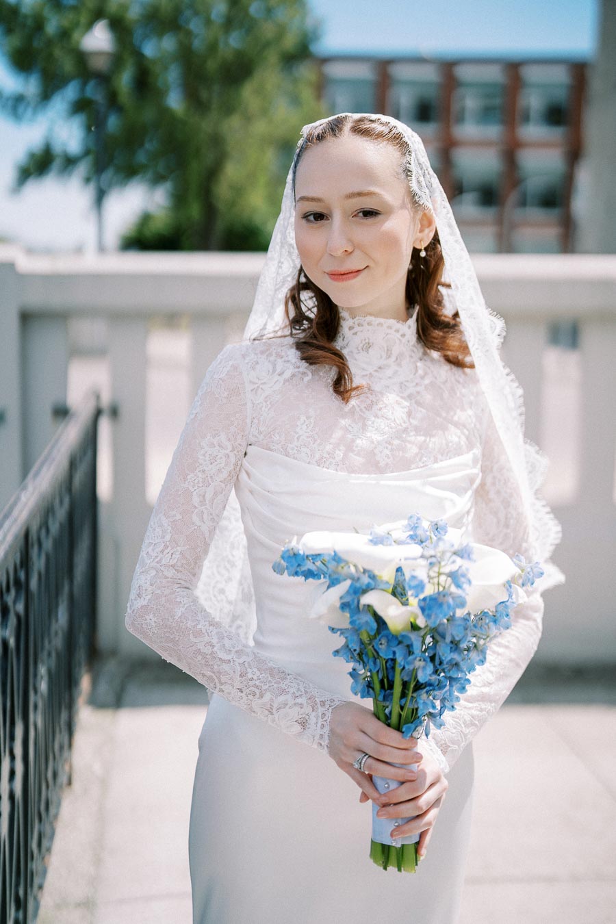 A bride in a white lace wedding dress and veil holding a bouquet of blue and white flowers, standing outdoors with a blurred background of trees and a building.