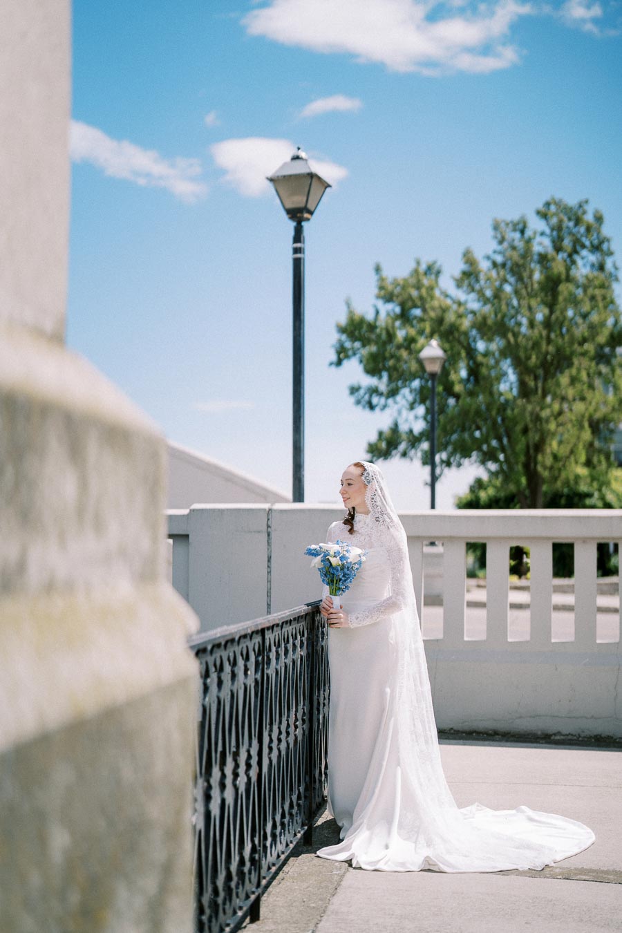 Bride in elegant white gown holding a bouquet of blue flowers on a sunny day, standing by an ornate iron railing with a streetlamp and trees in the background.
