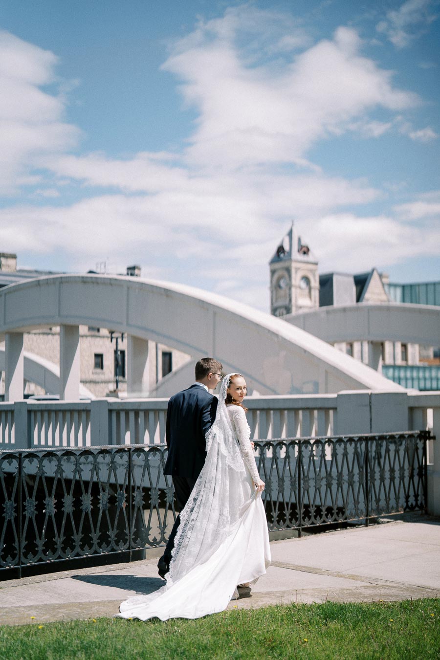 Wedding couple walking on a bridge under a bright blue sky, with bride in elegant white gown and groom in black suit, against a backdrop of historical architecture.