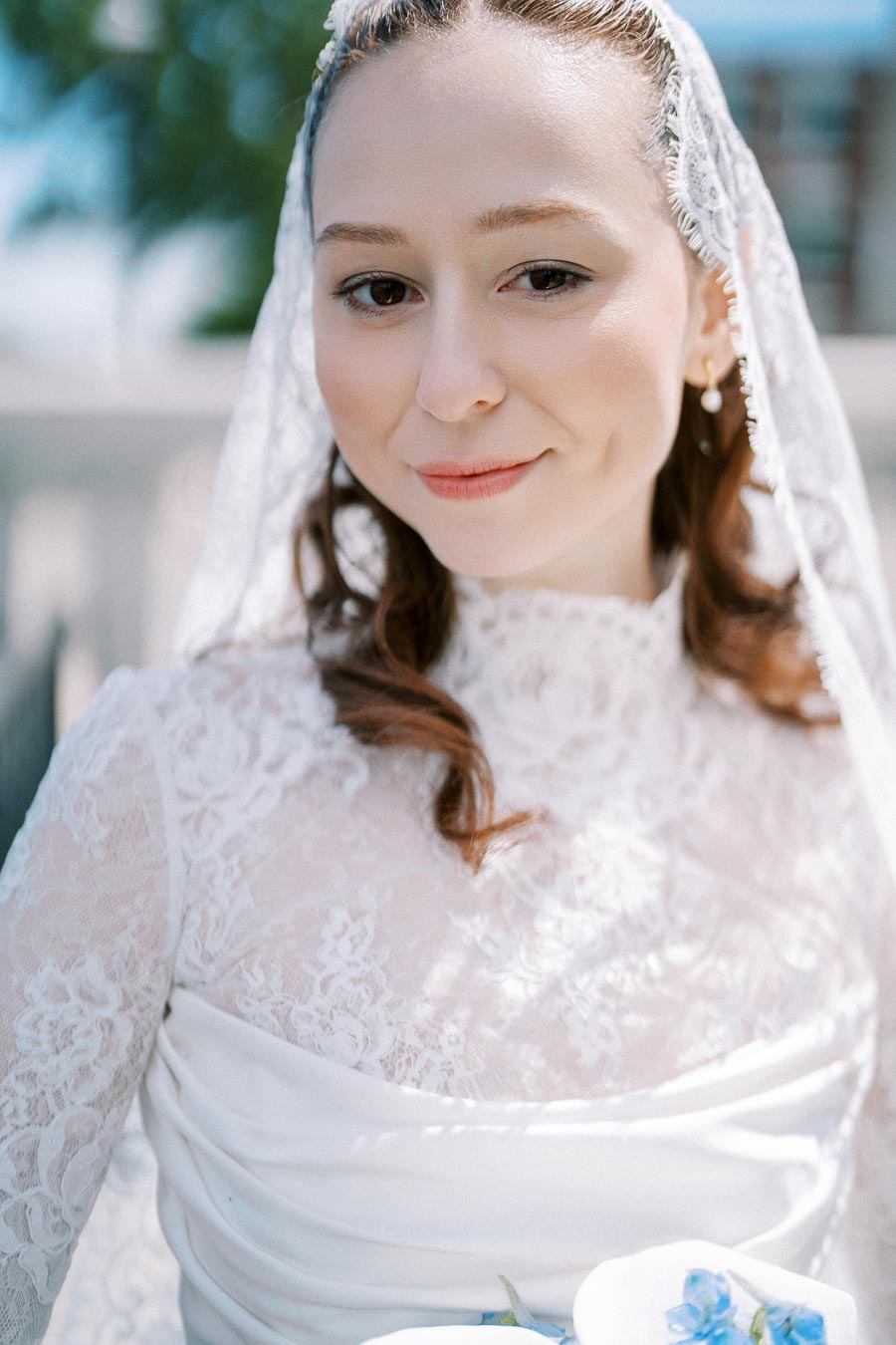 A bride in a lace wedding dress with a veil, smiling softly outdoors, holding a bouquet with blue flowers.