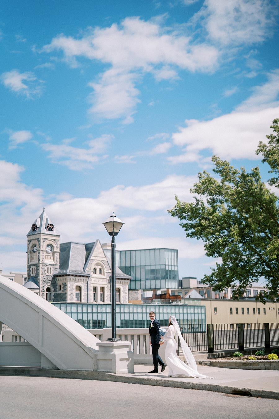 A bride in a flowing white gown and veil holds hands with a groom in a black suit while walking across a white bridge in a city. Behind them, historic and modern buildings contrast against a bright blue sky with scattered clouds. Trees and a black lamp post add to the scene's urban charm.
