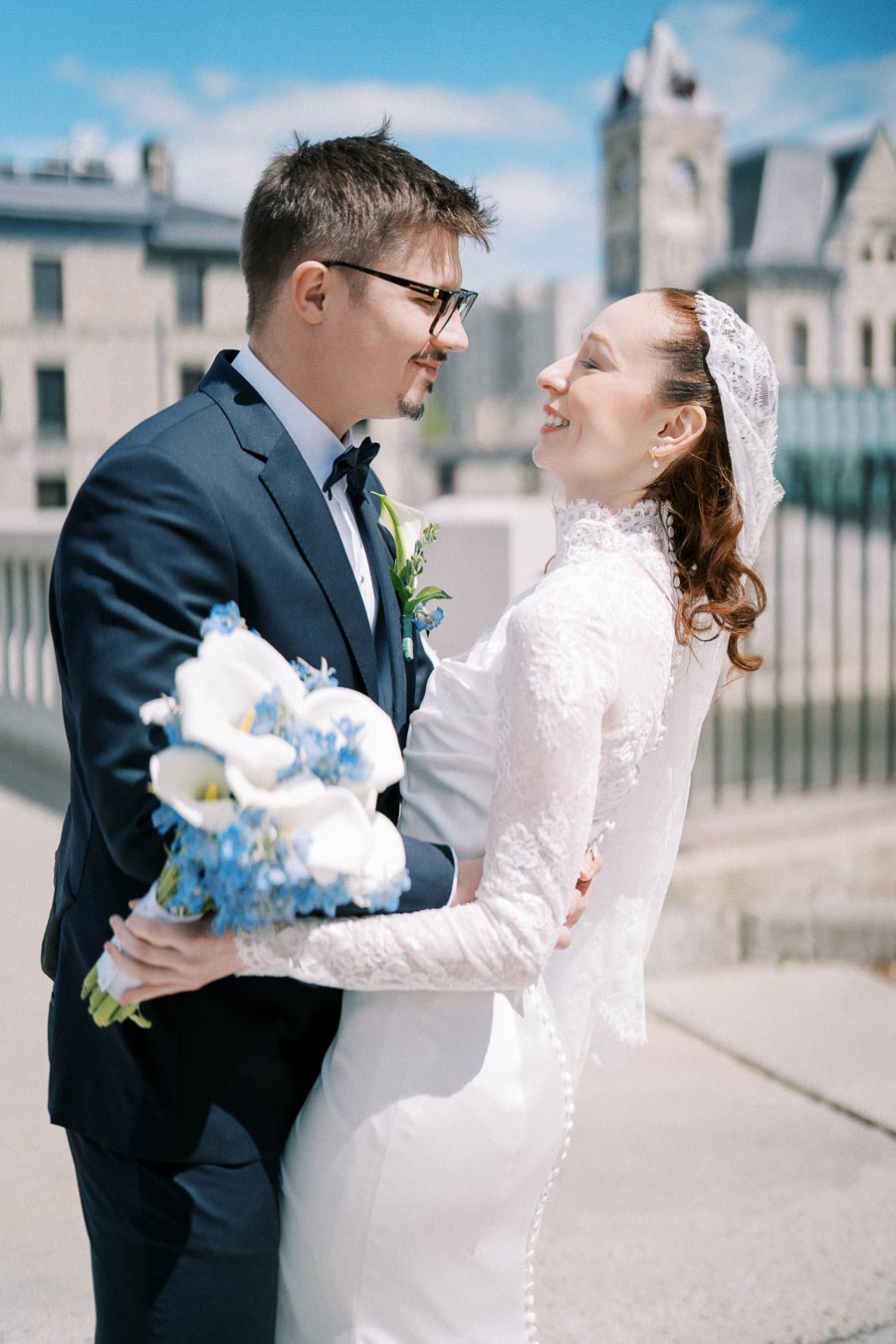 A bride and groom in a white wedding dress and dark suit share a joyful moment outside, holding a bouquet of white and blue flowers, with a historic building in the background.