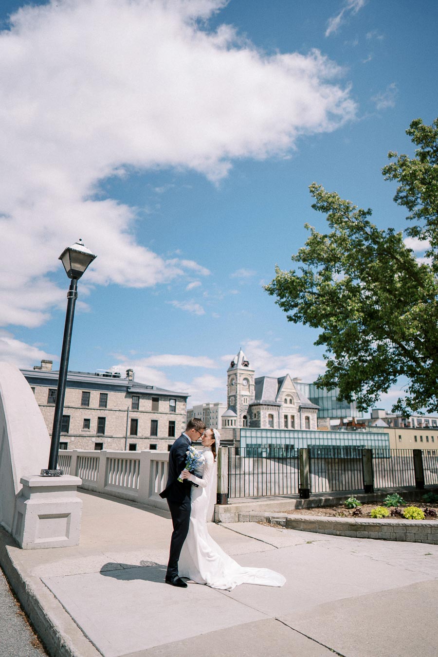 Elegant bride and groom sharing a kiss on a sunlit stone bridge, with historic architecture and a bright blue sky in the background, capturing a picturesque wedding moment.