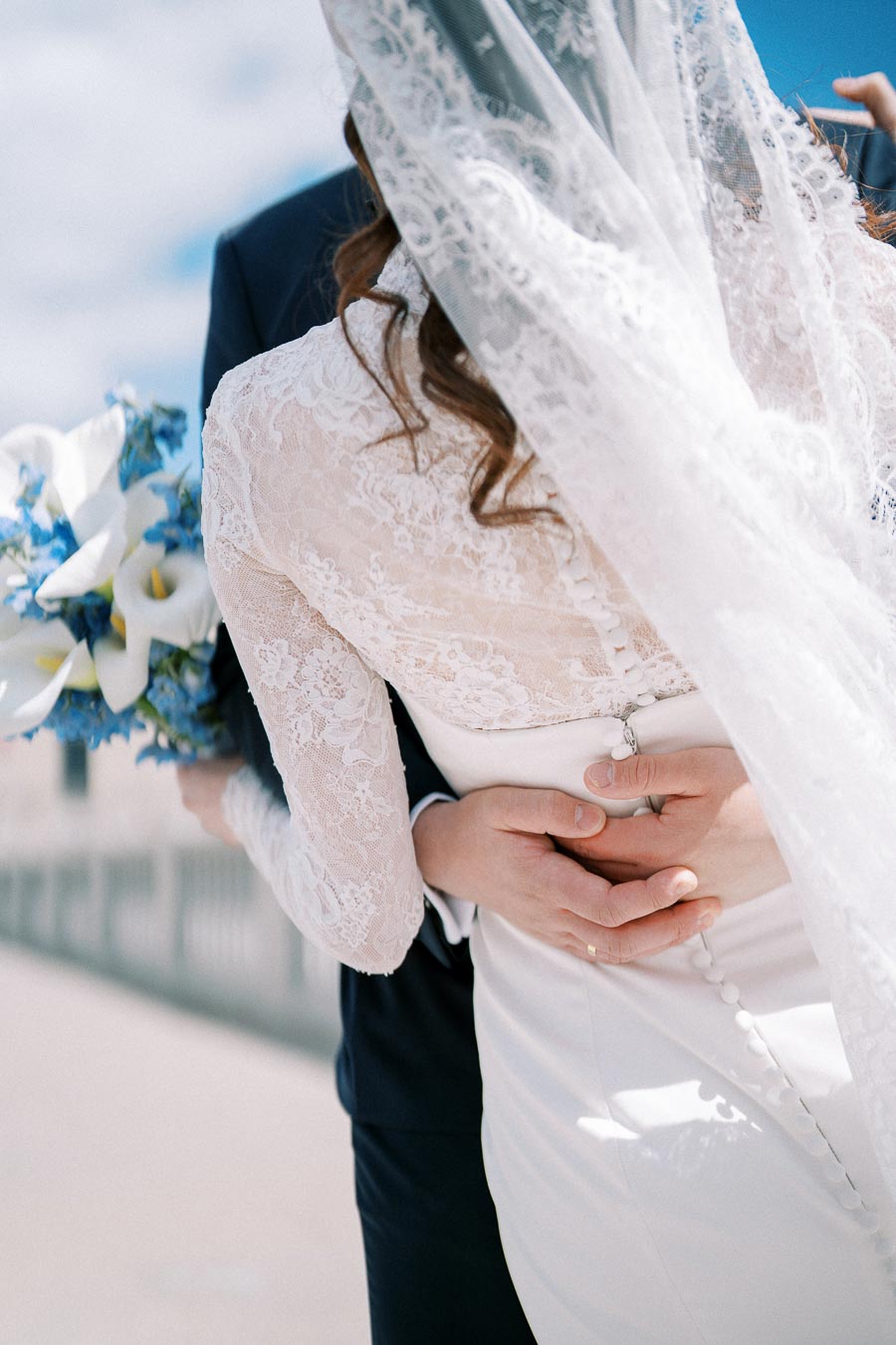 Elegant bride in lace wedding dress and veil holding blue and white floral bouquet, embraced by groom in dark suit, highlighting love and romance on a sunny wedding day.
