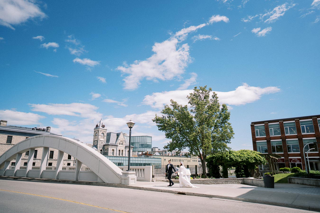 A bride and groom walking hand in hand on a sunny day, with a historic building and a modern structure visible in the background under a clear blue sky.