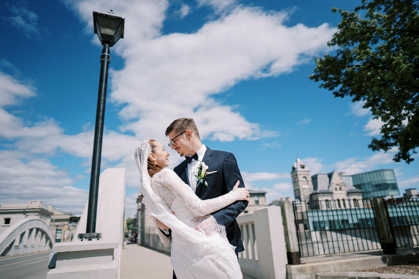 A bride and groom embracing joyfully on a bridge, under a clear blue sky, with elegant buildings in the background.
