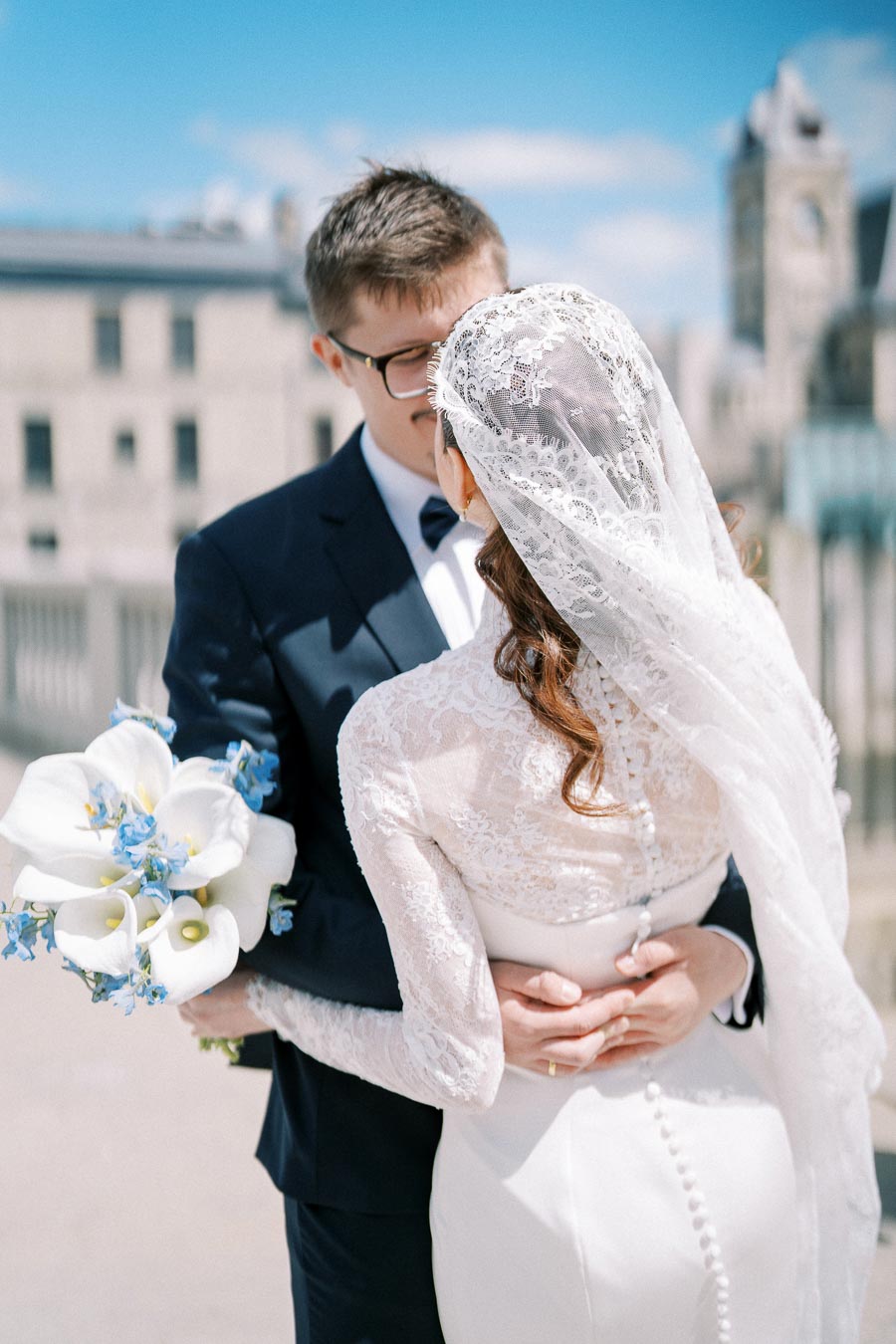 Elegant bride and groom embracing outdoors, bride in lace gown with bouquet of white and blue flowers, background of a historic cityscape under a clear blue sky.
