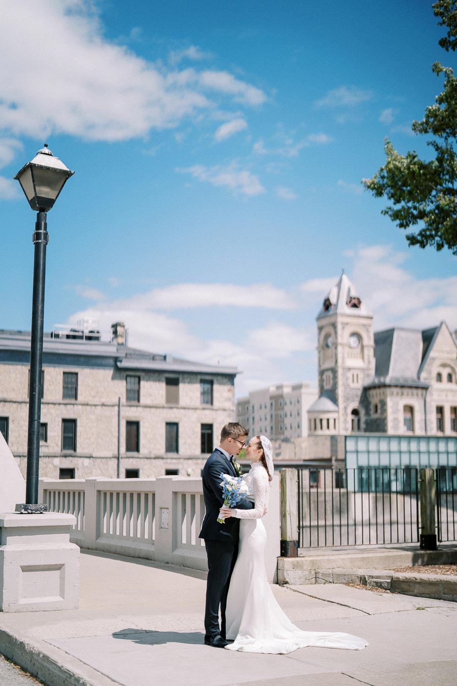 Wedding couple embracing on a city street with historic architecture in the background, featuring a bright blue sky and elegant bridal attire.