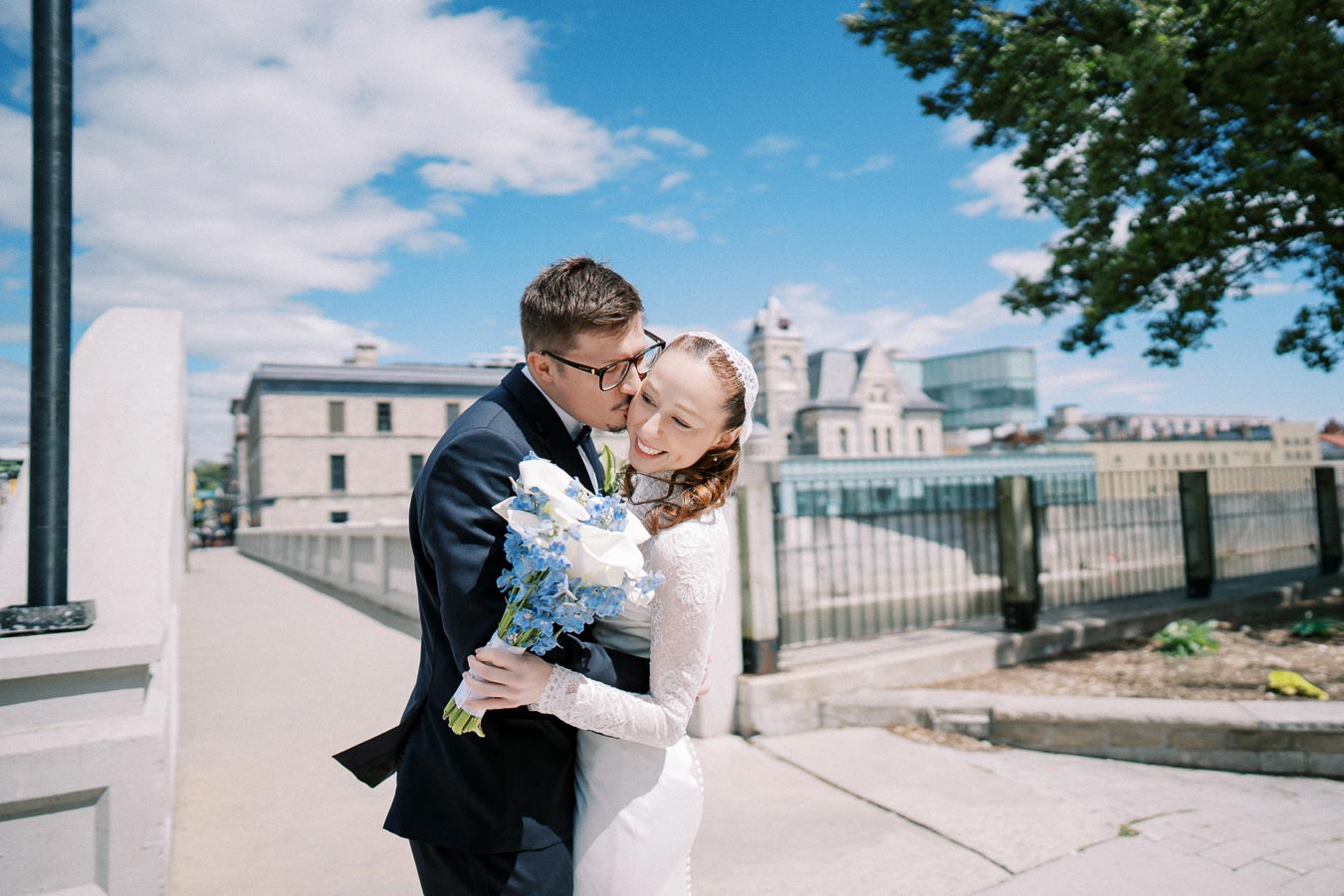 A joyful wedding couple embracing and smiling, with the bride holding a bouquet of white and blue flowers, set against a scenic urban backdrop on a sunny day.