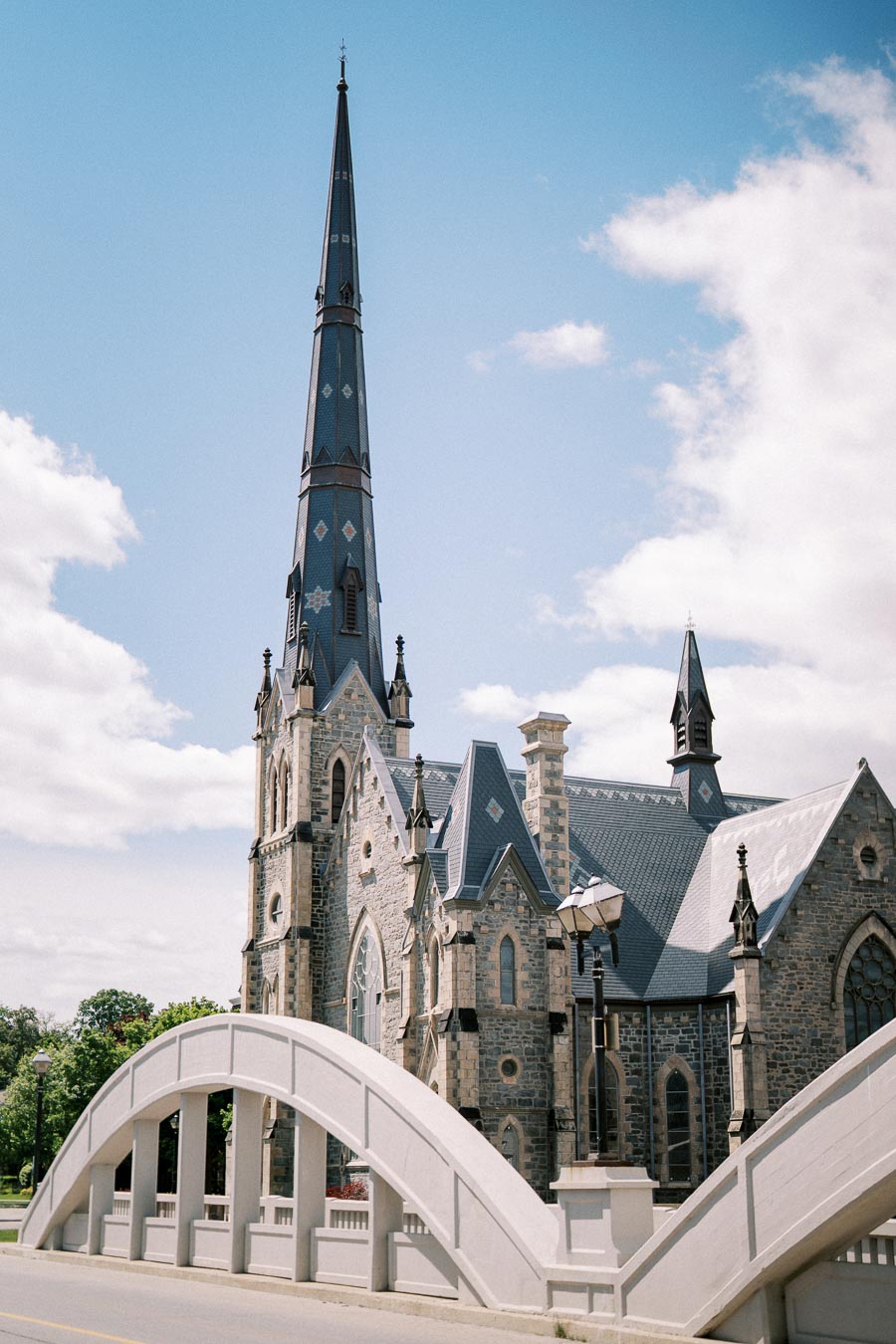 Gothic-style stone church with a tall spire under a clear blue sky, near a white arched bridge, surrounded by greenery.