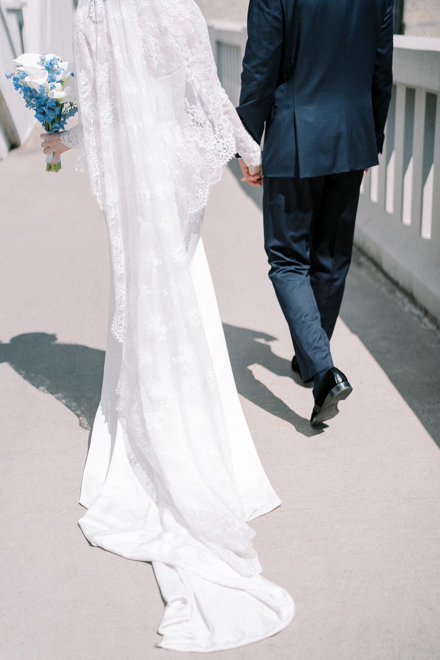 A bride and groom holding hands walking away, with the bride in a long white lace gown carrying a bouquet of white and blue flowers, and the groom in a dark suit, on a sunny day.