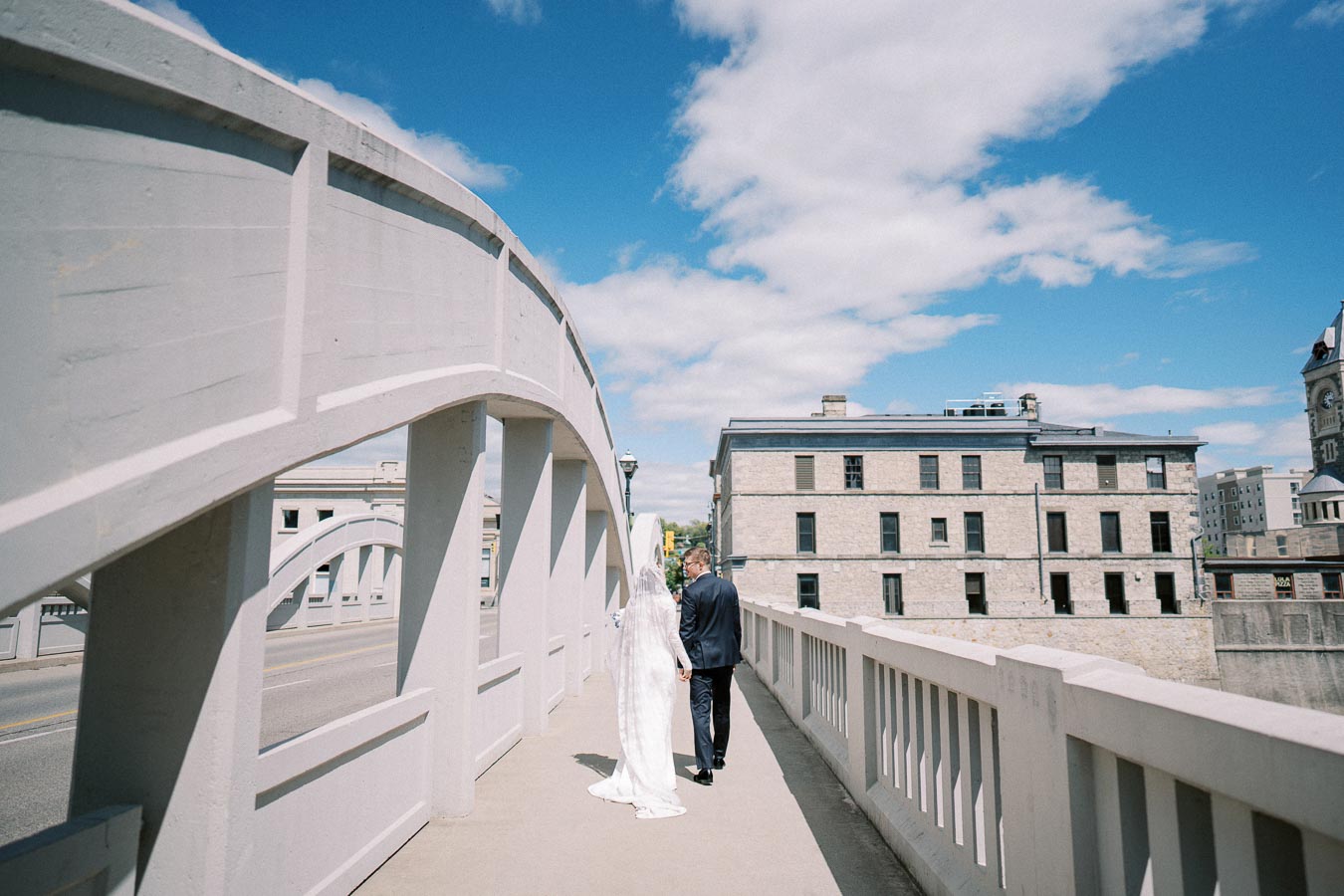 A bride and groom walking hand in hand on a historic urban bridge under a bright blue sky, with vintage stone buildings in the background.