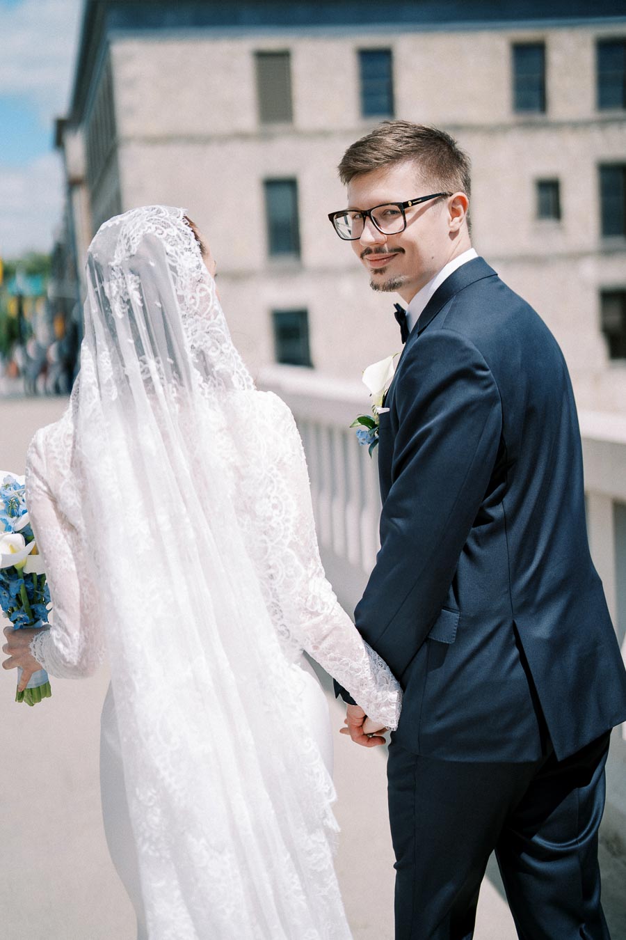 A bride in a lace wedding dress and veil holds hands with the groom in a dark suit and glasses, smiling while walking outdoors on a sunny day.