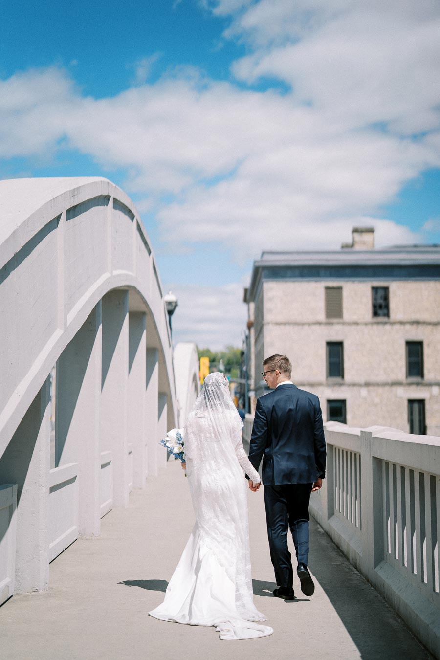 A newlywed couple holding hands while walking across a bridge, with the bride in a white wedding dress and veil, and the groom in a dark suit, under a clear blue sky.