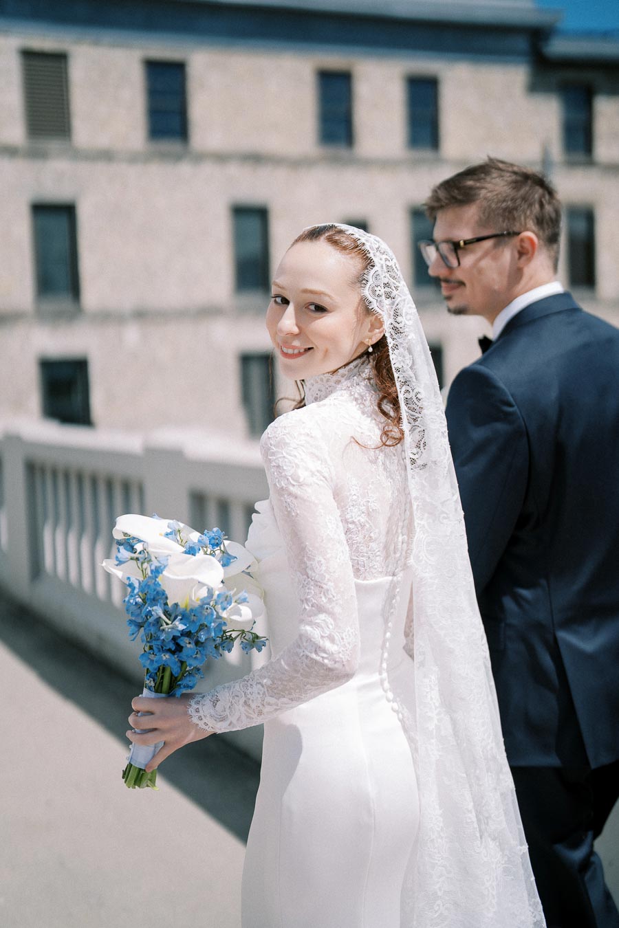 Elegant bride in lace gown holding a bouquet with blue and white flowers, standing next to groom on a sunny day outside, with a building in the background.