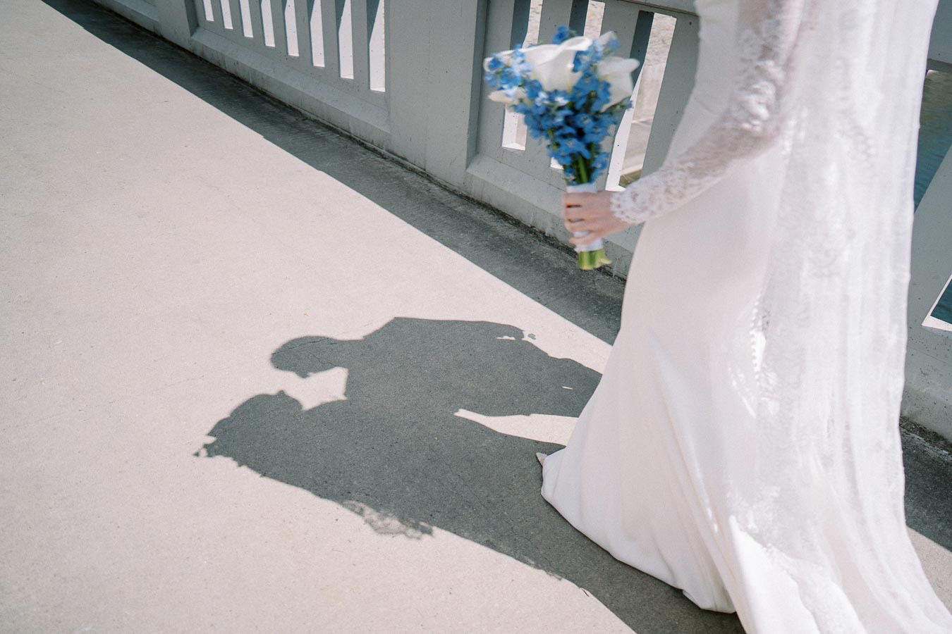 A bride in a white dress holding a bouquet of blue flowers walks across a bright bridge, casting a romantic shadow on the ground.