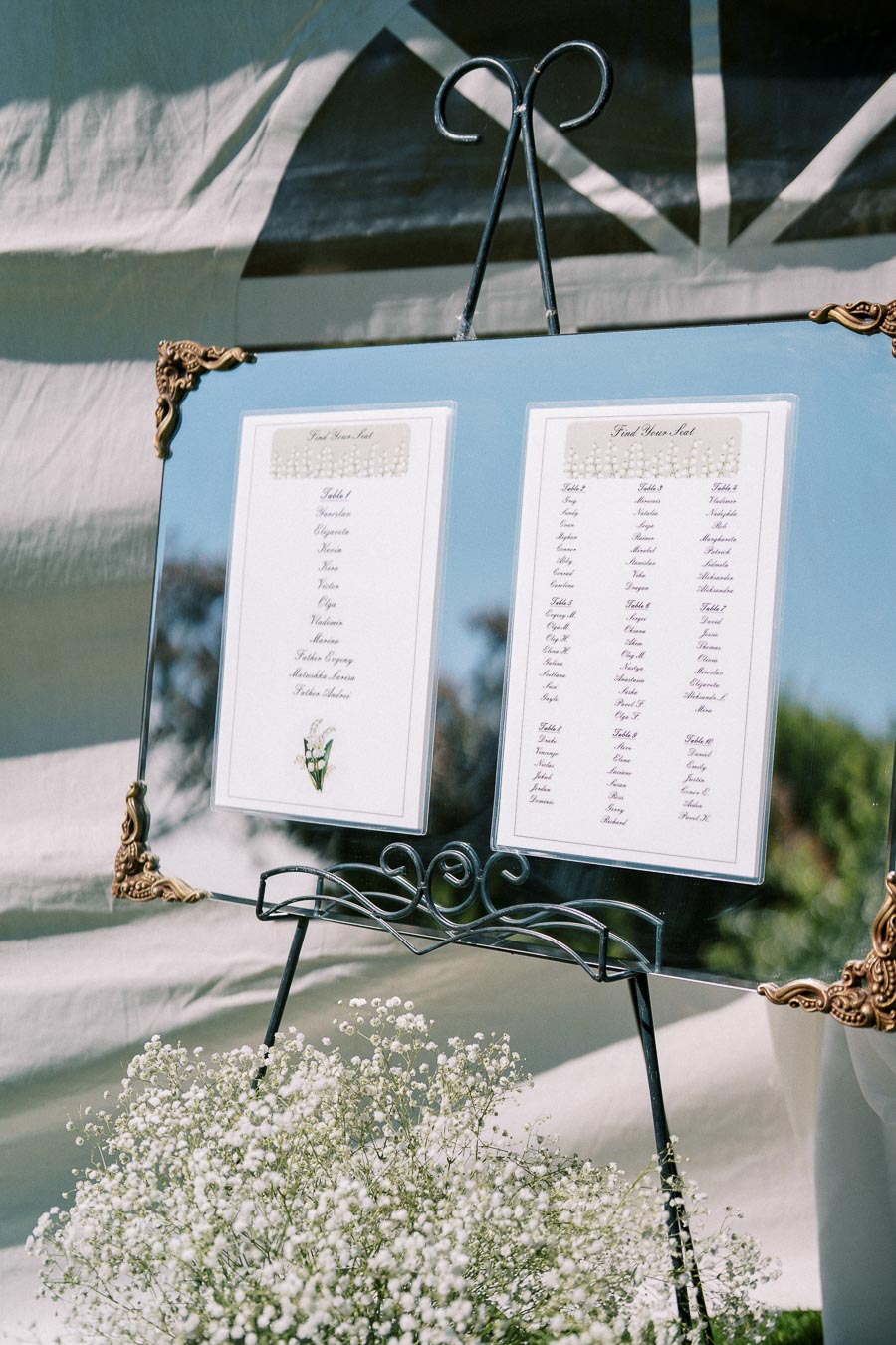 Wedding seating chart displayed on a decorative easel with ornate corners, featuring guest names under different table numbers. The elegant setup is surrounded by lush greenery and delicate baby's breath flowers, creating a sophisticated and welcoming atmosphere.