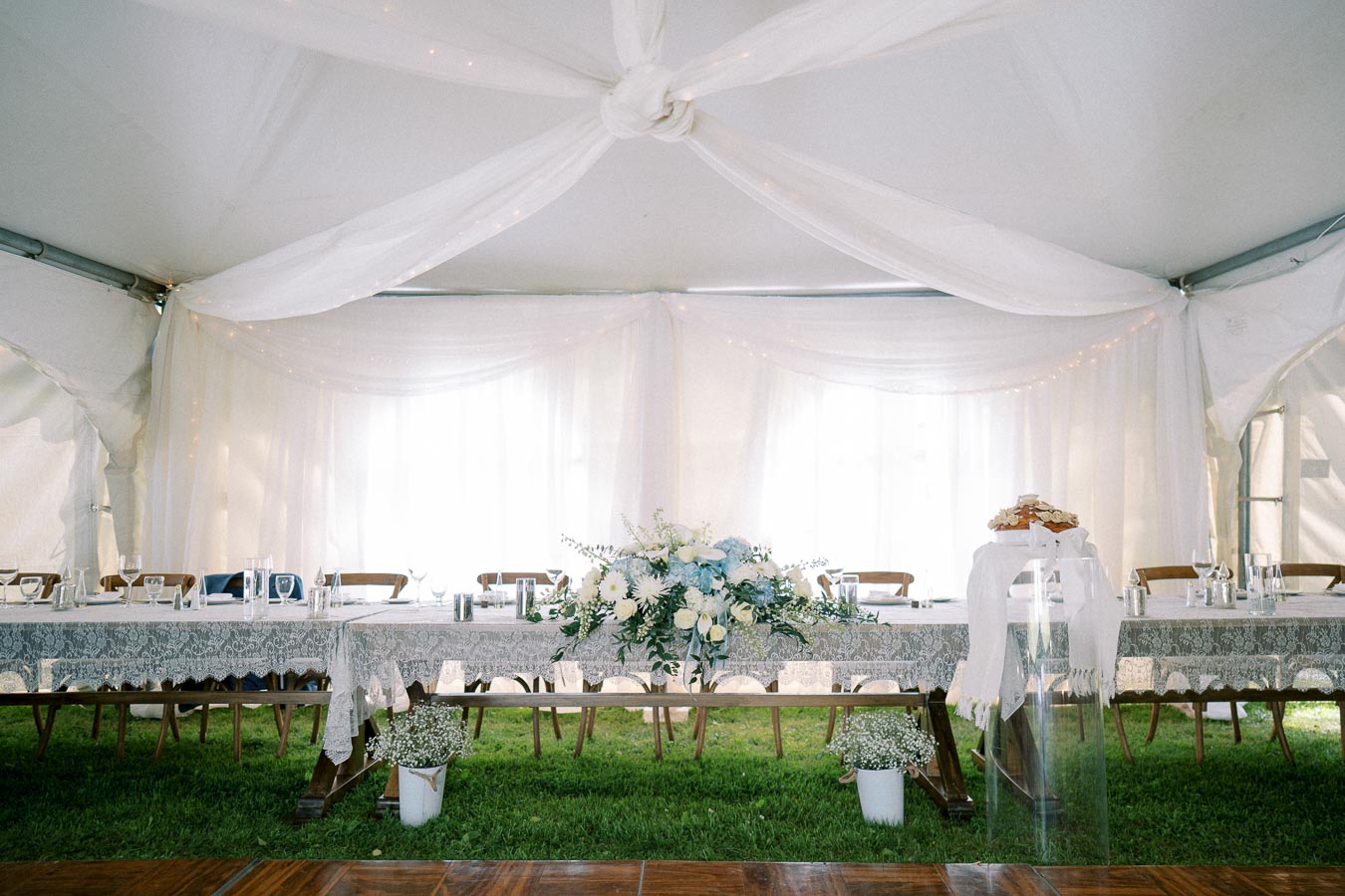 Elegant wedding reception setup under a white tent, featuring a beautifully decorated long table with lace tablecloths, floral centerpieces, and elegant glassware on a grassy floor.