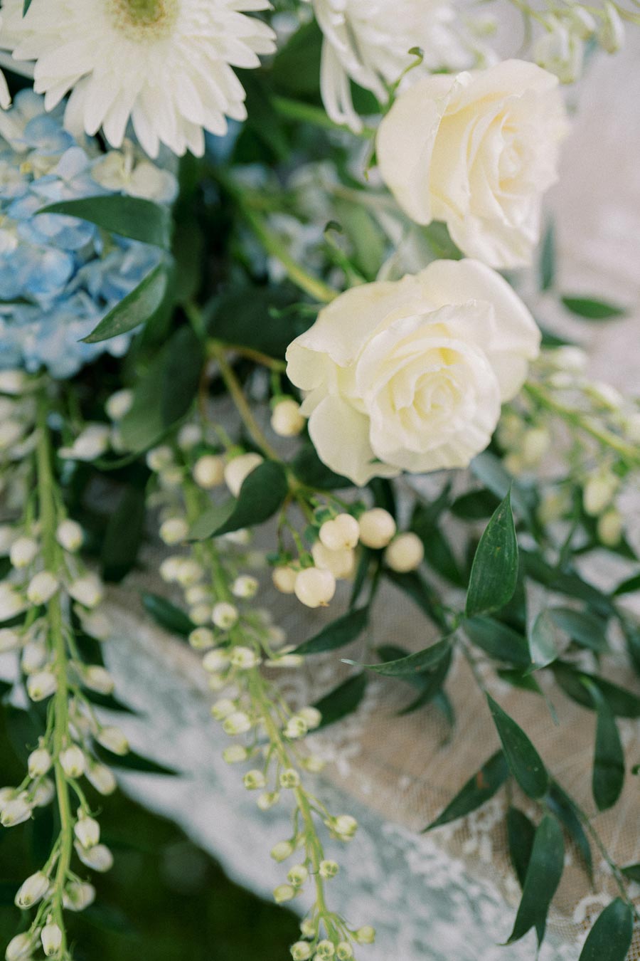 Elegant floral arrangement featuring white roses, daisies, blue hydrangeas, and greenery, perfect for a wedding or special event centerpiece.