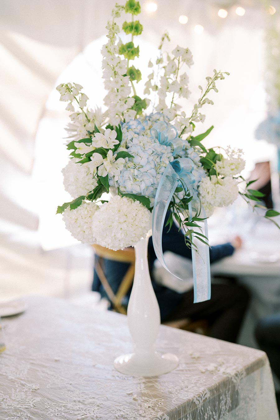 Elegant floral centerpiece with white and blue flowers in a white vase, displayed on a lace-covered table at a formal event.