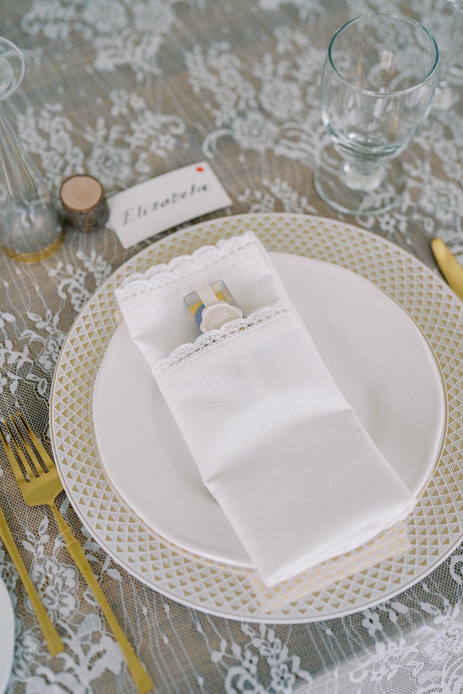 Elegant wedding table setting with a gold-trimmed plate and cutlery, lace tablecloth, neatly folded white napkin with a favor box, and a personalized name card reading Elizabeth.