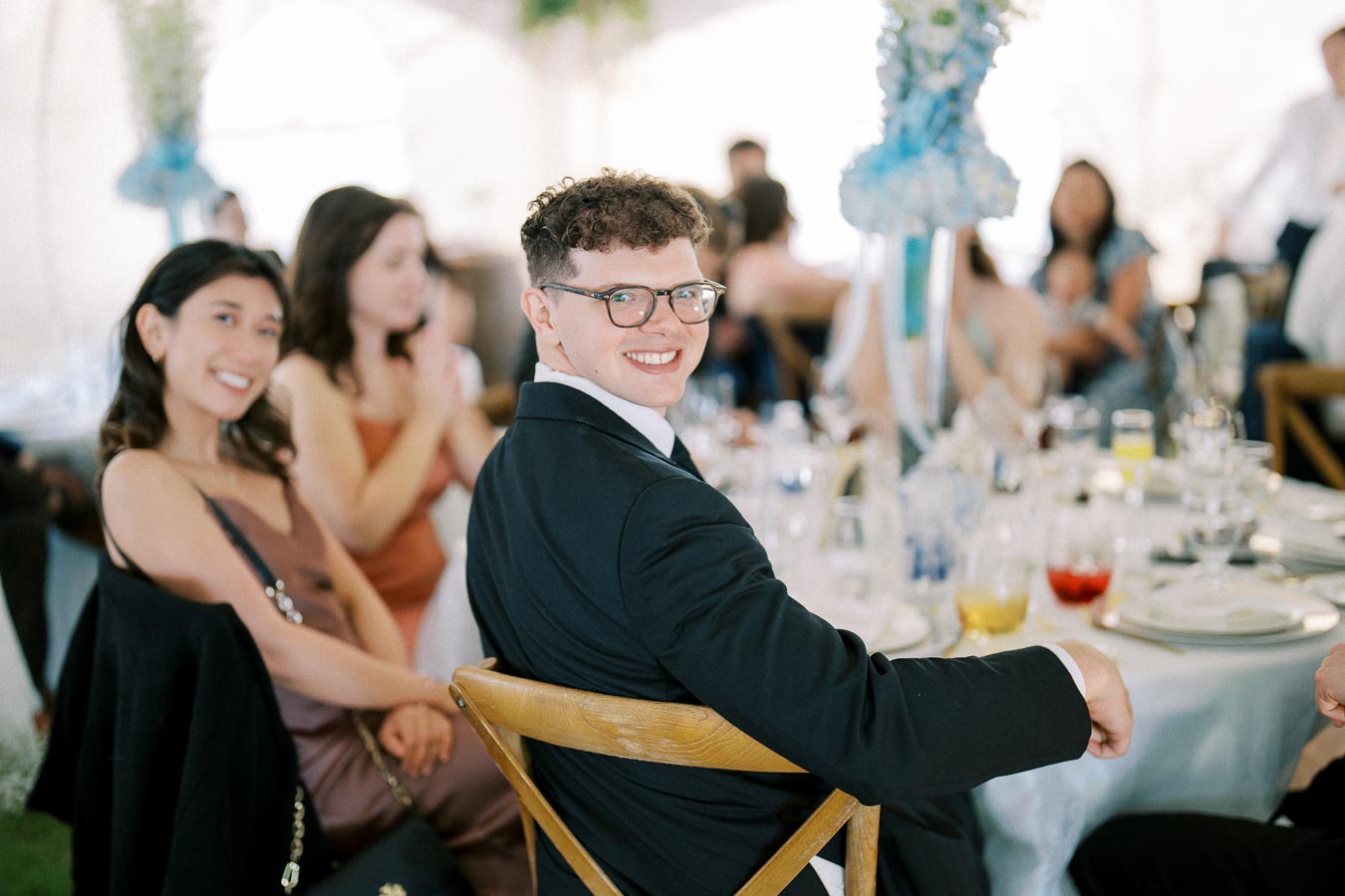 Smiling guest in formal attire at an elegant wedding reception, sitting at a beautifully set dinner table with other attendees in the background.