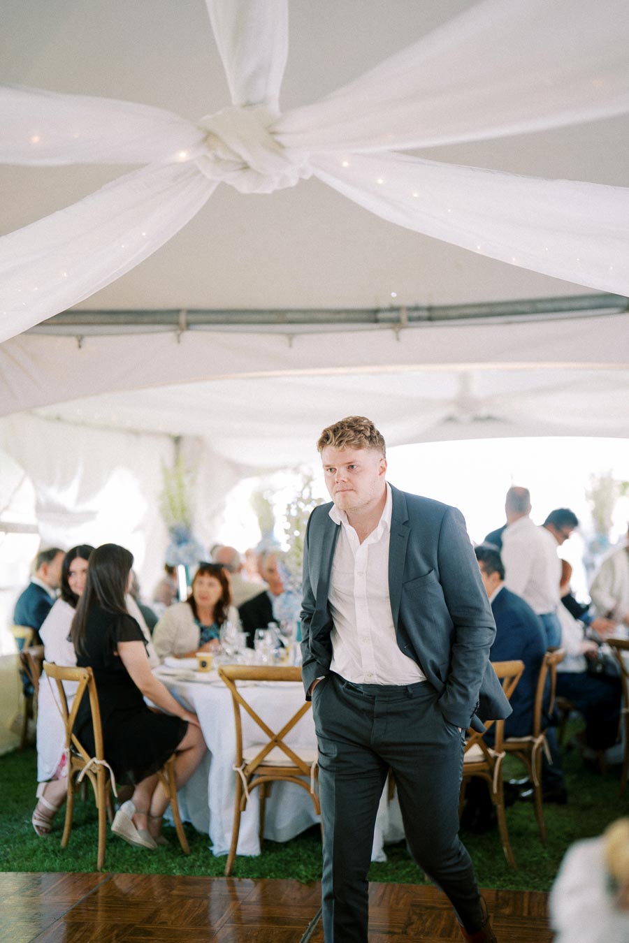Man in suit walking at an outdoor event under a white tent, with seated guests in the background.