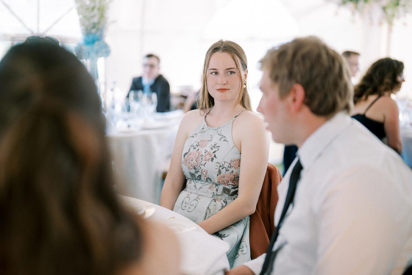 A woman in a floral dress sitting at a formal event, attentively listening to a conversation, with a blurred background of other guests and elegantly set tables.
