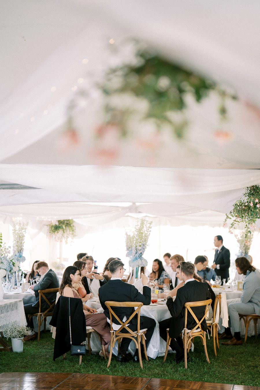 A group of elegantly dressed guests sitting at decorated tables inside a white tent at a wedding reception, with floral arrangements and ambient lighting.