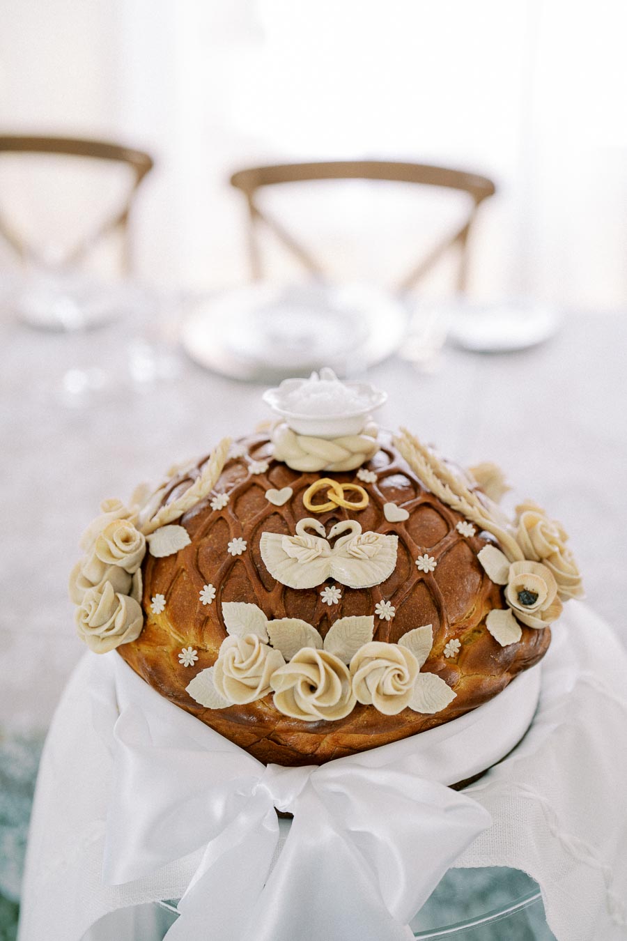 A beautifully decorated traditional bread, adorned with intricate dough designs including flowers and leaves, symbolizing celebration, placed on a table with an elegant white cloth, perfect for weddings or festive events.