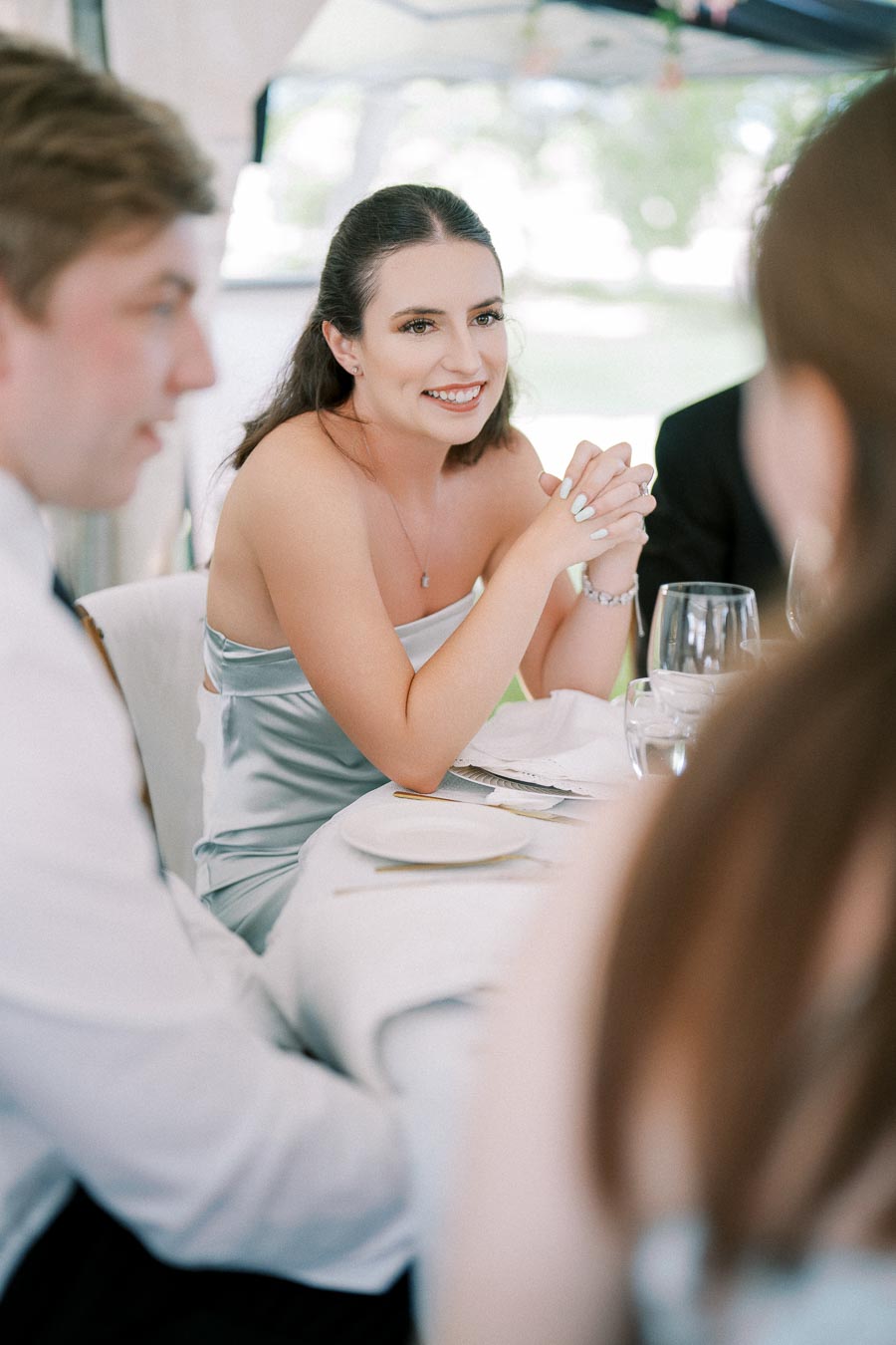 Young woman in elegant light blue dress smiling and conversing at a formal dining event with other guests, soft natural light streaming in from the window.