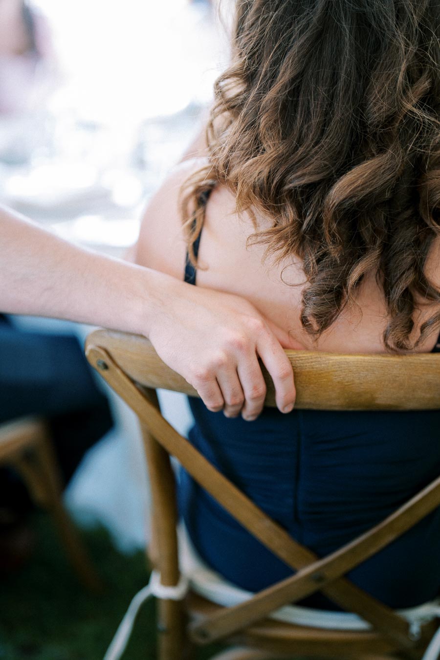 A man gently rests his hand on the back of a woman's chair, highlighting a moment of connection and support, with her curly hair flowing over her dark dress.
