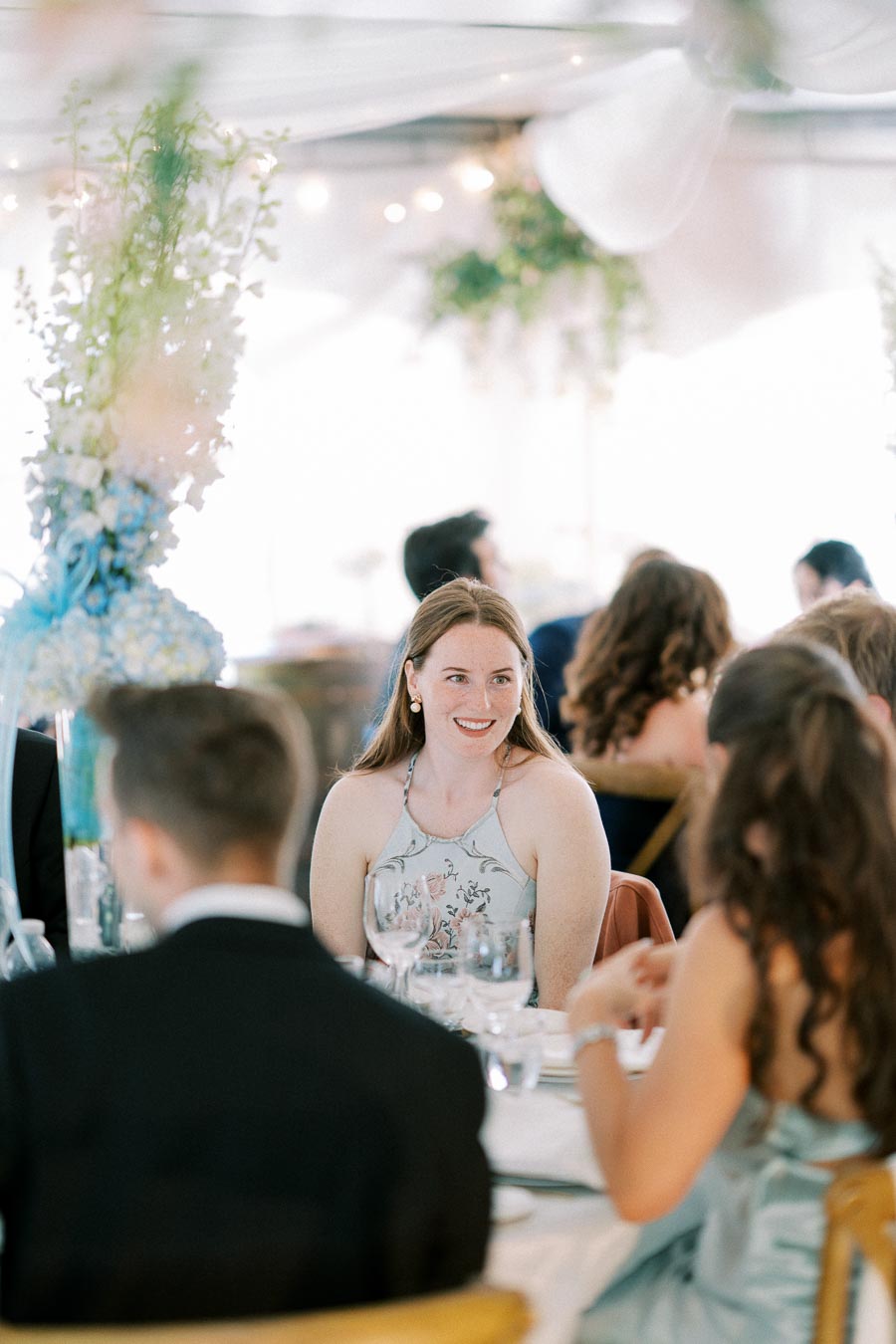 Smiling woman in floral dress seated at a formal dinner table, surrounded by other guests in elegant attire, with decorative floral arrangements in the background.