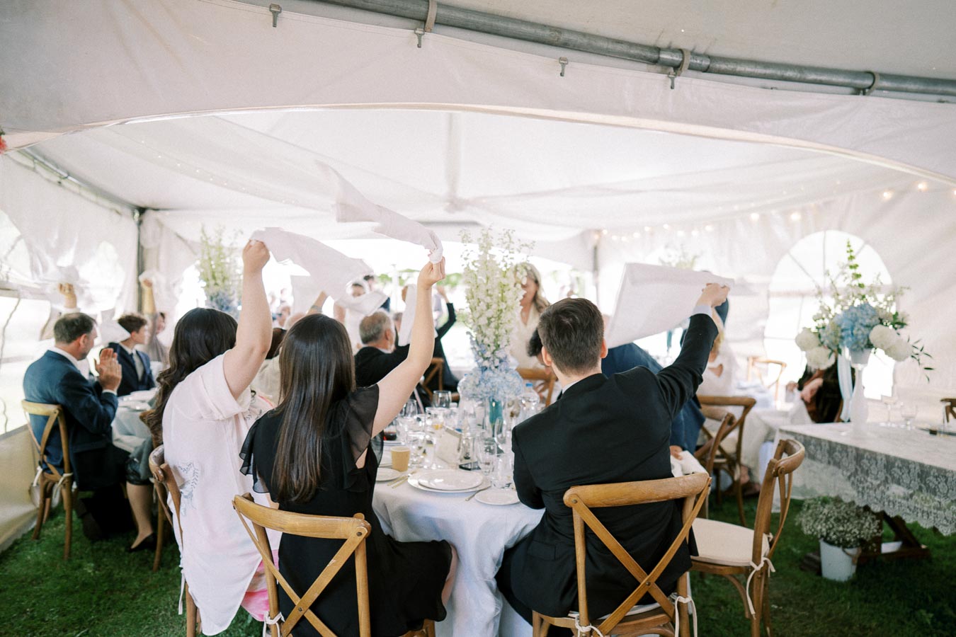 A lively wedding reception in a tent, with guests seated around tables waving napkins in celebration, surrounded by elegant floral centerpieces and decor.