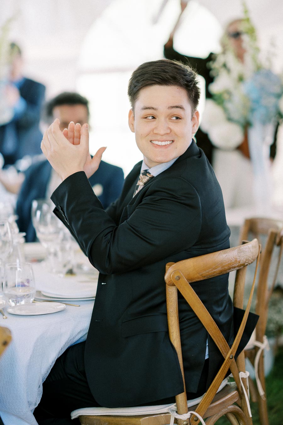 A smiling man in a suit clapping at a formal event, seated at a table with elegant tableware and chairs, with blurred guests and floral decorations in the background.