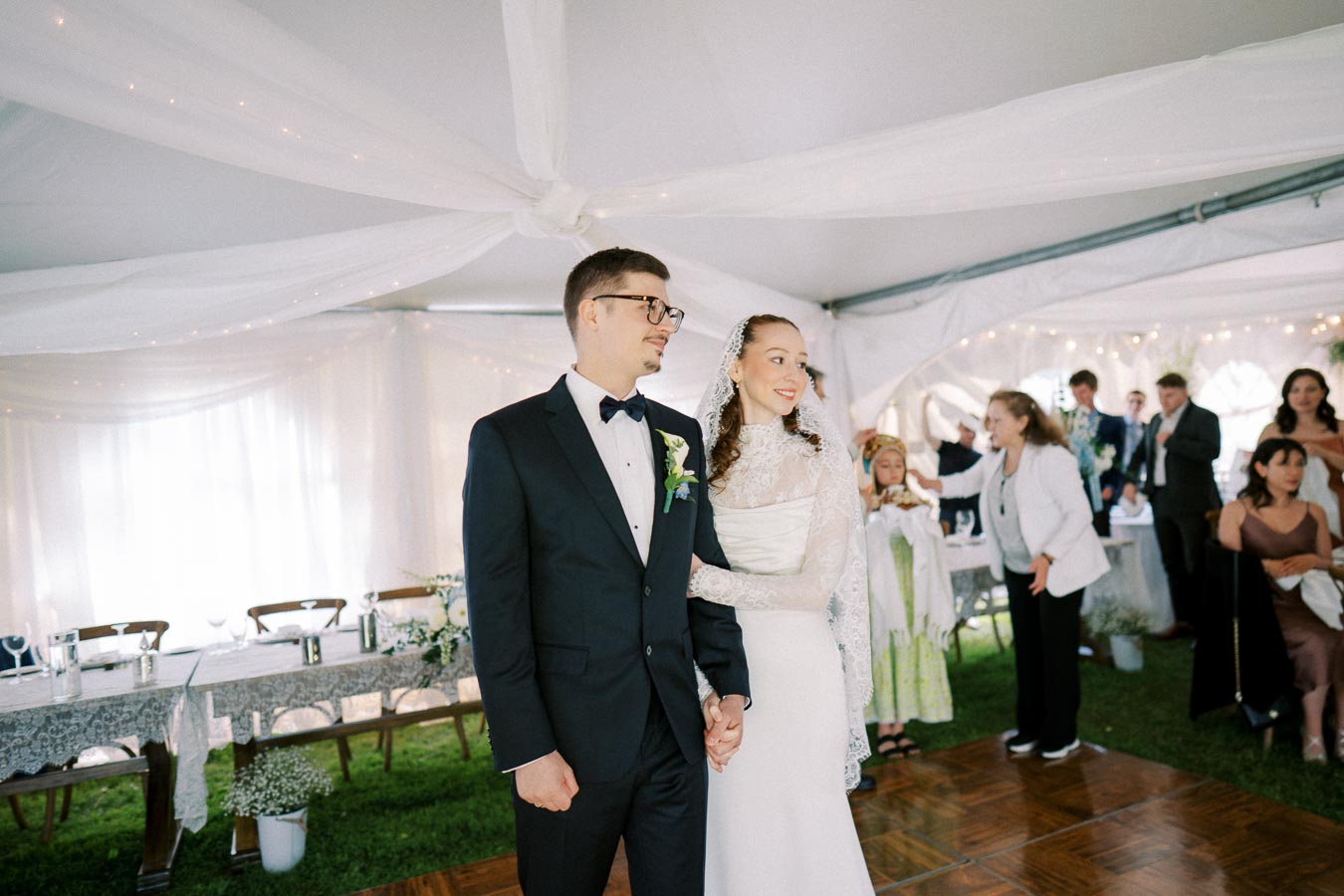 A bride and groom stand holding hands under a decorated tent at their wedding reception, surrounded by guests.