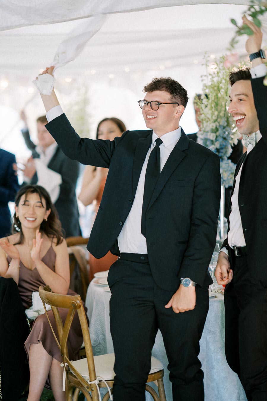 A group of well-dressed wedding guests joyfully waving napkins and clapping in celebration under an elegant white tent.