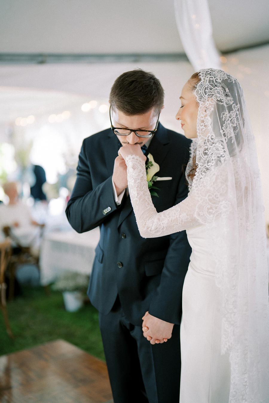 A groom tenderly kisses his bride's hand during an intimate wedding ceremony, with the bride wearing an elegant lace wedding dress and veil, creating a romantic and emotional moment.